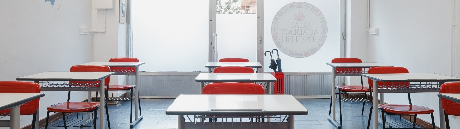 Aula de The Real Academy Of English con pupitres blancos, sillas rojas y puerta acristalada en una academia de inglés